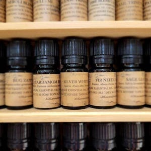 May include: A shelf of small brown glass bottles with black caps, each labeled with a different essential oil name and description. The labels are white with black text. The oils include Bug Zap, Cardamom, Silver White, Fir Needle, Sage, and Led MI.