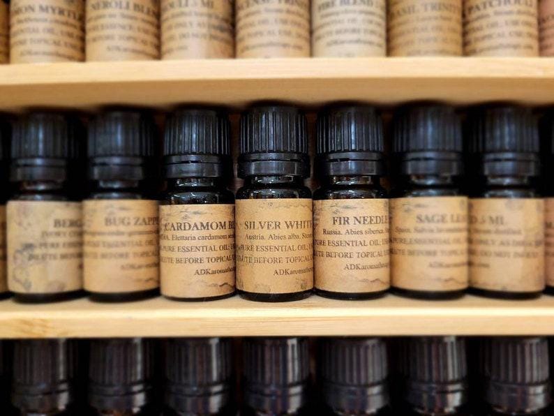 May include: A shelf of small brown glass bottles with black caps, each labeled with a different essential oil name and description. The labels are white with black text. The oils include Bug Zap, Cardamom, Silver White, Fir Needle, Sage, and Led MI.