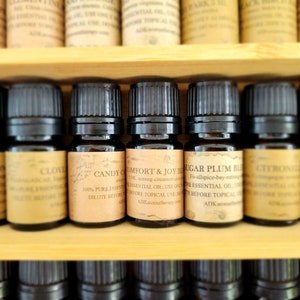 May include: A wooden shelf with rows of small brown glass bottles filled with essential oils. The bottles have labels with names like "Lemon", "Clove", "Candy Comfort", "Sugar Plum", "Citron", and "Rosemary".