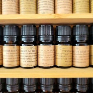 May include: A close-up of a wooden shelf with rows of small, dark brown glass bottles filled with essential oils. The bottles have labels with text that includes the names of the oils, such as "Douglas Fir", "Cinnamon Leaf", and "Citronella".
