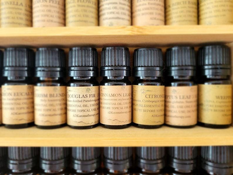 May include: A close-up of a wooden shelf with rows of small, dark brown glass bottles filled with essential oils. The bottles have labels with text that includes the names of the oils, such as "Douglas Fir", "Cinnamon Leaf", and "Citronella".