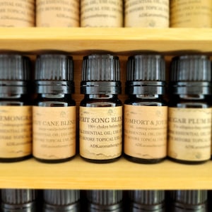 May include: A close-up of a wooden shelf with rows of small brown glass bottles filled with essential oils. The bottles have labels with names like "Lemongrass", "Body Cane Blend", "Spirit Song Blend", "Comfort & Joy", "Sugar Plum", and "Citron".