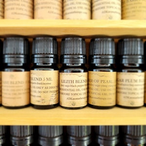 May include: A close-up of a shelf with ten small brown glass bottles filled with essential oils. The bottles are labeled with names like "Douglas Fir", "Lilith Blend", and "Sugar Plum". Each bottle contains 5 ml of oil.