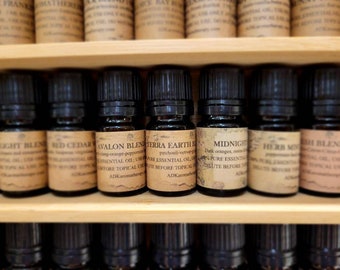 May include: A wooden shelf with rows of small, dark brown glass bottles with black caps. Each bottle has a label with a different name and description of the essential oil inside. The labels are all in a vintage style with brown paper and white text. The names of the oils include "Avalon Blend", "Terra Earth", "Midnight", "Herb Mint", and "Light Blend".