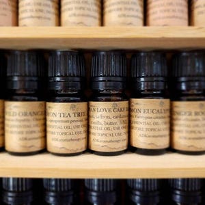May include: A close-up view of a shelf with small glass bottles filled with essential oils. The bottles have brown labels with white text and are arranged in a row. The labels include names like "Lemon Tea Tree", "Vanilla Love Cake", and "Lemon Eucalyptus".
