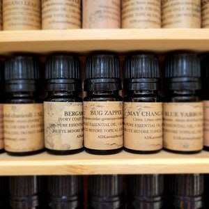 May include: A shelf of small, dark brown glass bottles with labels. The labels are cream-colored with black text. The bottles are filled with essential oils. The labels read: Cypress Needle, Chavicoll 5 ml, Bergamot Ivory Coast, Bug Zapper, May Change, Blue Yarrow, Marjoram.