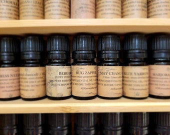 May include: A shelf of small, dark brown glass bottles with labels. The labels are cream-colored with black text. The bottles are filled with essential oils. The labels read: Cypress Needle, Chavicoll 5 ml, Bergamot Ivory Coast, Bug Zapper, May Change, Blue Yarrow, Marjoram.