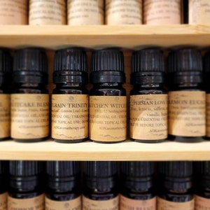 May include: A shelf of small glass bottles filled with essential oils. The bottles are labeled with names like "Grain Trinity", "Kitchen Witch", and "Persian Love".