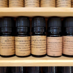 May include: A shelf of small brown glass bottles with black caps, each labeled with a different essential oil blend. The labels include names like "Lavender Ice Blend", "Aqua Flores", "Dragons Blood", and "Mermaid Blend".