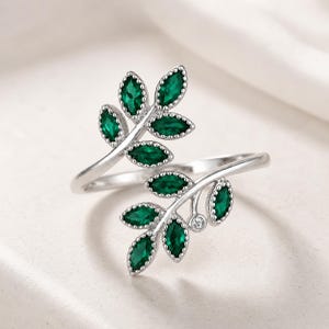 May include: A silver ring with a leaf design. The ring features two leaf branches adorned with green, oval-shaped gemstones. A small, clear gemstone adds detail. The ring is set against a soft, white background.