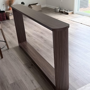 May include: A long, narrow console table with a dark brown wood grain finish. The table features a flat top surface and a matching bottom shelf, supported by two vertical, semi-circular pillars with a distinct vertical fluted texture.
