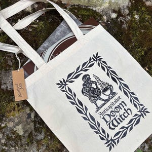 Könnte beinhalten: Eine cremefarbene Stofftasche mit einer schwarzen Tintenillustration einer Figur neben einem brennenden Kessel, umrahmt von einer Blattbordüre. Der Text lautet "Anticapitalist Doom Witch". Ein braunes Pappetikett mit der Aufschrift "Westford Mill" ist befestigt.