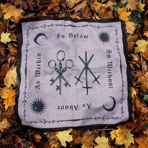 Op de afbeelding: Een vierkante stoffen doek met een zwarte kanten rand met een bedrukt ontwerp van drie sleutels en drie zwaarden. De tekst luidt As Within, So Without, As Above, So Below. Het ontwerp bevat zwarte zon- en maaniconen op een lichtgrijze achtergrond.