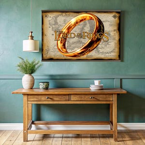 Puede incluir: Un póster enmarcado con el logotipo de El Señor de los Anillos y un anillo dorado. El póster está sobre un fondo de mapa. El póster está colgado sobre una mesa de madera con dos cajones. Un jarrón con vegetación y una taza y platillo pequeños están sobre la mesa.