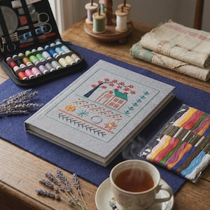 May include: A gray book with a cross-stitch design of a house, tree, and floral elements. A black sewing kit, spools of thread, and a packet of colorful embroidery floss are also visible. A cup of tea and lavender sprigs complete the scene.