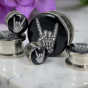 May include: A collection of black and silver ear plugs with a rock and roll hand gesture design. The plugs feature a glossy black surface with a white skeleton hand graphic. The plugs are made of metal and are displayed on a white surface.