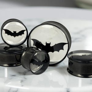 May include: Black and white bat-themed ear gauges in various sizes. The gauges feature a black bat silhouette against a white background. The plugs are made of a glossy black material.