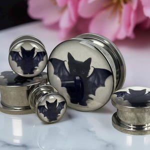 May include: A collection of silver-toned ear gauges with a black bat design against a cream background. The bat has outstretched wings. The gauges are displayed on a white surface with pink flowers in the background.