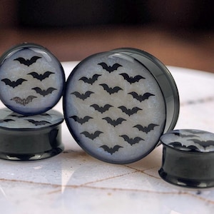 May include: A set of three circular ear plugs with black rims and a translucent white, glittery interior. Each plug features a repeating pattern of small, black bat silhouettes printed across the surface, creating a consistent dark motif.