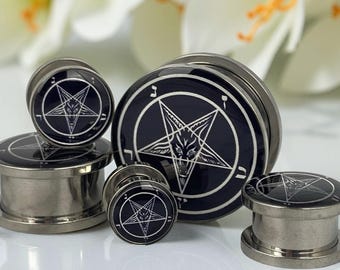 May include: A collection of dark gray metal ear plugs with a black and white pentagram design. The plugs vary in size and are displayed against a blurred background of white flowers. The pentagram is a five-pointed star within a circle.