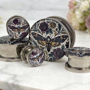May include: A collection of stainless steel ear plugs with a decorative design. The plugs feature a moth and floral pattern in shades of black, brown, purple, and cream. The moth has a skull on its body. The plugs are in various sizes.
