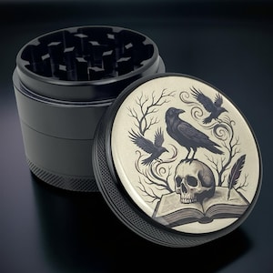 May include: Black herb grinder with a detailed graphic on the lid. The lid features a skull, ravens, and a book design in black and cream colors. The grinder is made of metal and has a textured grip.