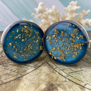 May include: A pair of ear gauges with a dark teal background and gold leaf flakes. The gauges have a clear, rounded edge. The background is a gradient of teal and blue. The gauges are displayed on a map.
