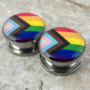 May include: A pair of stainless steel ear gauges with a rainbow pride flag design. The flag includes the colors of the transgender pride flag, the black and brown stripes of the progress pride flag, and the traditional rainbow pride flag colors.