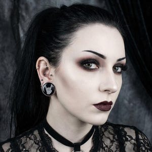 May include: A woman with dark hair and dramatic makeup, wearing a black lace top and a choker. A large black ear gauge with a white hand gesture design is visible in her ear. The overall aesthetic is gothic and alternative.