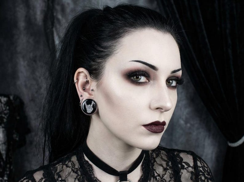 May include: A woman with dark hair and dramatic makeup, wearing a black lace top and a choker. A large black ear gauge with a white hand gesture design is visible in her ear. The overall aesthetic is gothic and alternative.
