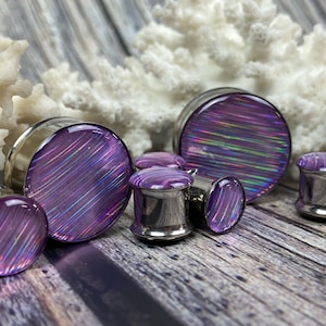 May include: A set of six iridescent purple ear gauges with a silver metal base. The gauges have a striped pattern and are arranged on a wooden surface.