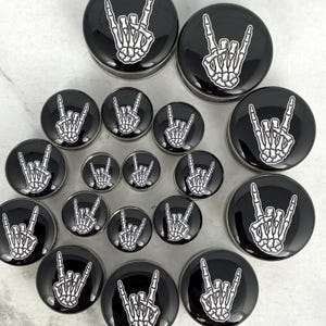 May include: A collection of black and silver ear gauges featuring a white skeleton hand making the rock and roll hand gesture. The gauges vary in size and are arranged on a light surface.