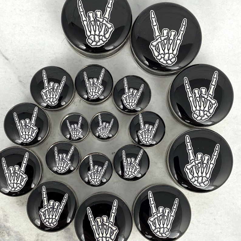 May include: A collection of black and silver ear gauges featuring a white skeleton hand making the rock and roll hand gesture. The gauges vary in size and are arranged on a light surface.