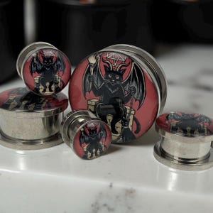 May include: A collection of stainless steel ear gauges with a red and black design. The design features a black cat with bat wings, holding a broom and a chain, set against a red background. The gauges vary in size.