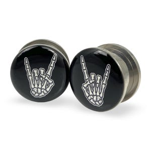 May include: Pair of silver-toned metal ear gauges with black circular faces. Each face features a white, stylized illustration of a skeleton hand making the rock and roll hand gesture. The gauges are designed for stretched ear piercings.