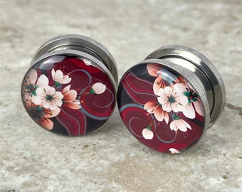 May include: A pair of silver ear gauges with a red and black floral design. The design features white flowers with green leaves on a dark red background.