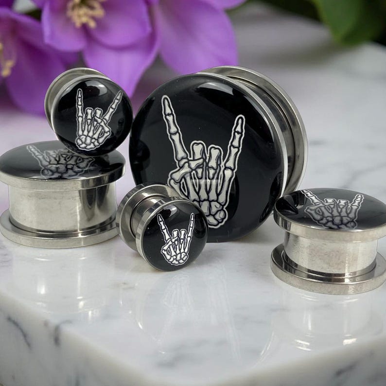 May include: A collection of black and silver ear plugs with a rock and roll hand gesture design. The plugs feature a glossy black surface with a white skeleton hand graphic. The plugs are made of metal and are displayed on a white surface.