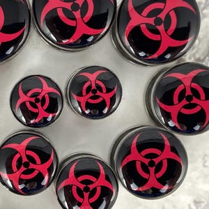 May include: Set of eight black and red biohazard symbol ear gauges. The gauges are made of metal and have a shiny finish.
