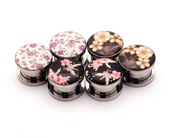 May include: Six pairs of stainless steel ear gauges with floral designs. The gauges feature a variety of floral patterns in shades of pink, white, and brown.