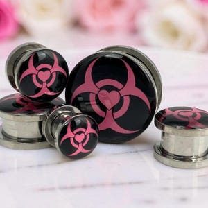 May include: A collection of stainless steel ear gauges with a black and pink biohazard symbol design. The symbol includes a heart shape in the center. The gauges are in various sizes and are displayed on a white surface.