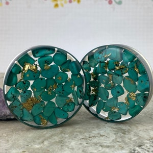 May include: A pair of teal green and gold ear gauges with a clear resin base. The gauges are filled with small teal green stones and gold flakes.