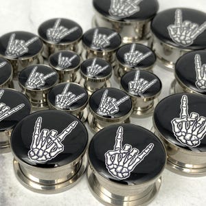 May include: A collection of stainless steel ear gauges with black tops featuring a white skeleton hand in a rock-and-roll gesture. The gauges vary in size and are displayed on a white surface.