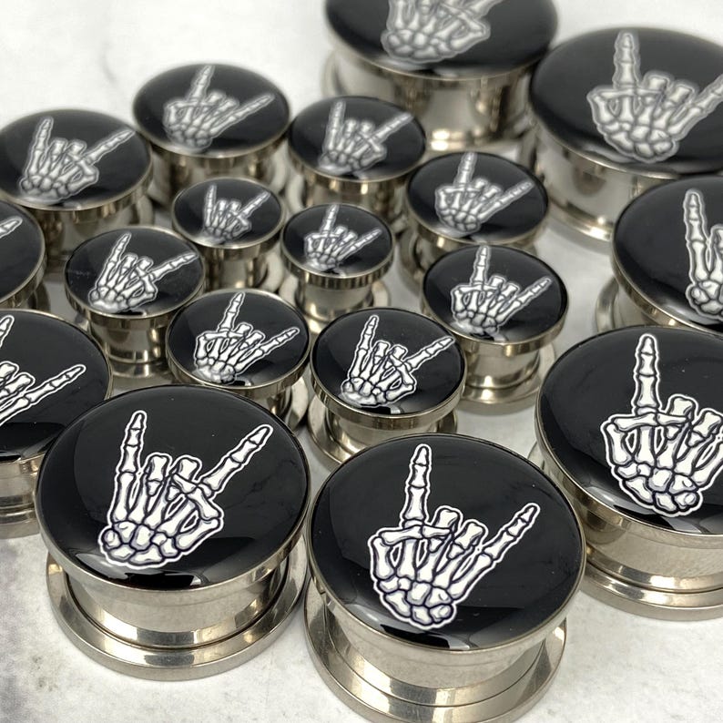May include: A collection of stainless steel ear gauges with black tops featuring a white skeleton hand in a rock-and-roll gesture. The gauges vary in size and are displayed on a white surface.