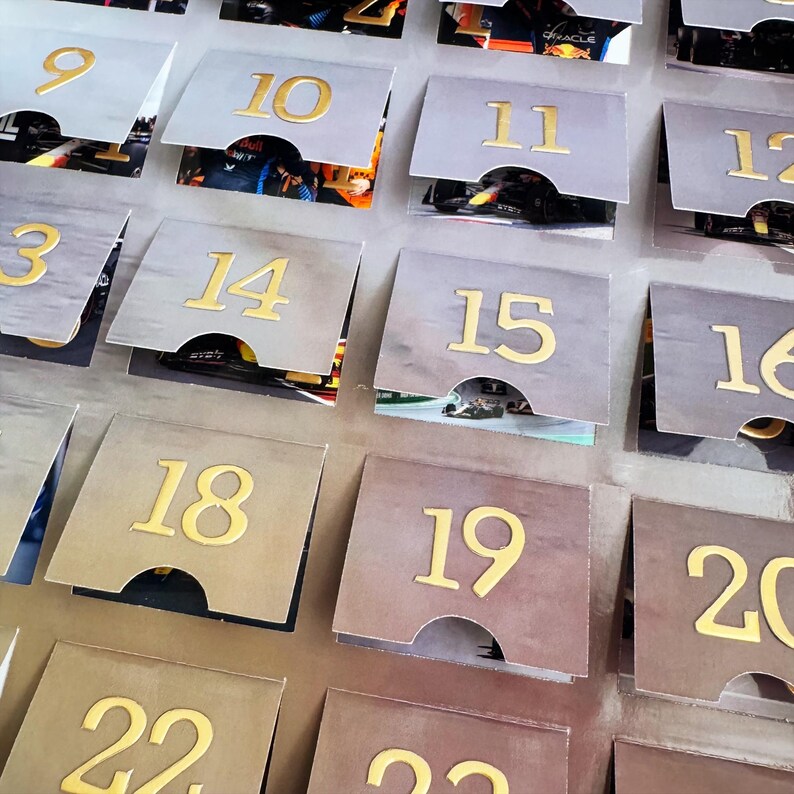 Puede incluir: Un primer plano de un calendario con los n&uacute;meros del 9 al 22 impresos en dorado sobre un fondo gris. Cada n&uacute;mero est&aacute; en una solapa separada que se puede levantar para revelar una imagen debajo.
