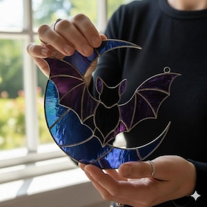 Puede incluir: Atrapasoles de vidriera con un murciélago y una luna creciente en tonos azules, morados y negros. El murciélago está centrado en la luna, con las alas extendidas. El atrapasoles tiene un marco de metal y un lazo para colgar. Un artículo decorativo para Halloween o decoración gótica.