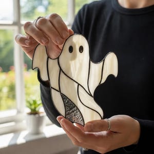 Puede incluir: Un atrapasoles de vitral con forma de fantasma, compuesto por segmentos de vidrio blanco opaco unidos por líneas de soldadura oscuras. El fantasma tiene dos ojos circulares negros y un panel de vidrio texturizado en la parte inferior.