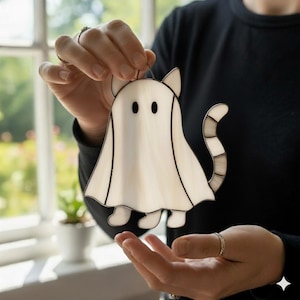 May include: A stained glass cat-shaped ghost decoration. The white ghost has black eyes and a striped tail. The cat ghost has a black outline and is held up by a thin wire loop.