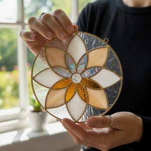 May include: A circular stained glass suncatcher featuring a geometric flower pattern with twelve petals. The design incorporates textured clear, iridescent, and amber-colored glass segments held together by thin gold-toned metal solder lines.