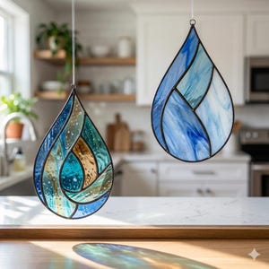 May include: Two stained glass teardrop ornaments with black metal frames hang against a blurred kitchen backdrop. One features blue, teal, and gold hues, while the other showcases shades of blue and white. Both are suspended by thin white cords.