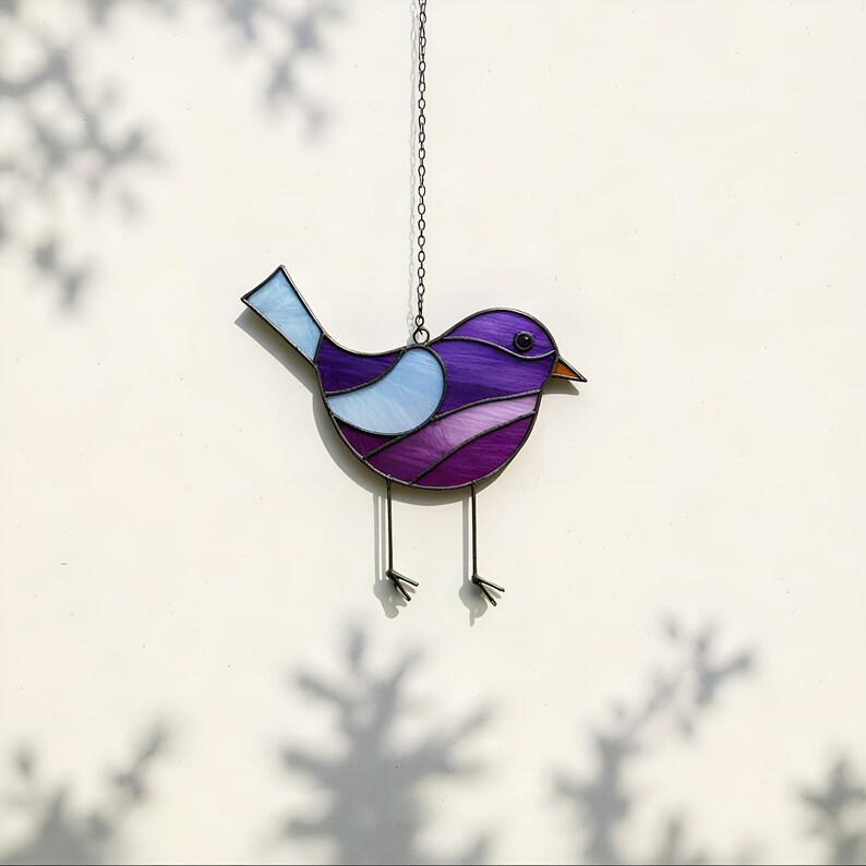 May include: Purple and blue stained glass bird hanging from a chain. The bird has a long tail and a small beak.